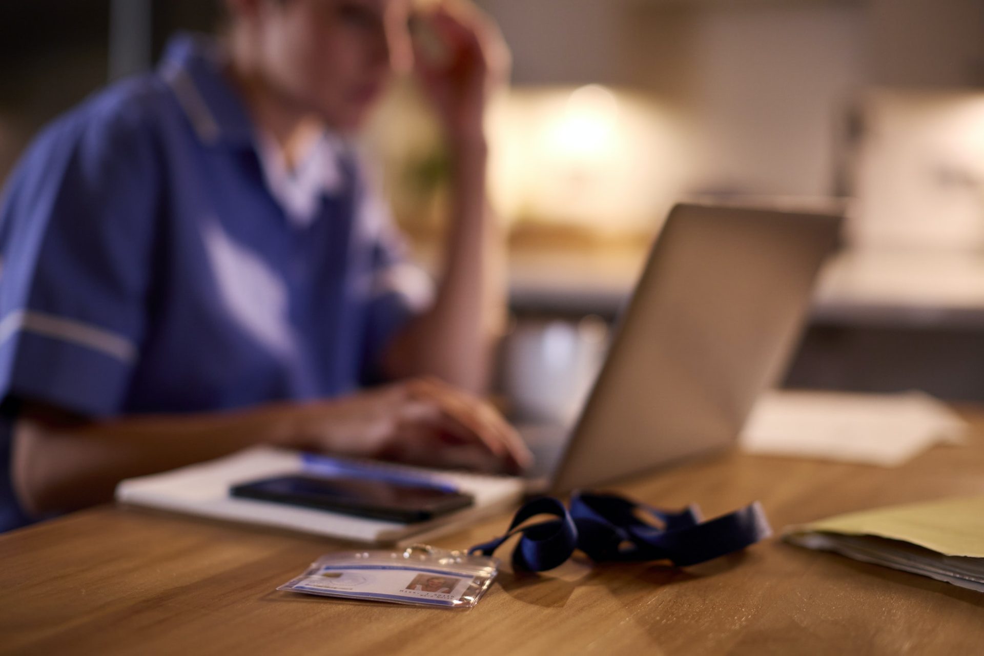 Woman In Medical Uniform Working Or Studying On Laptop At Home At Night With Security ID Lanyard